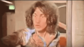 Man with curly hair wearing a shiny shirt mimics aiming a gun and shooting in a slow, exaggerated manner.