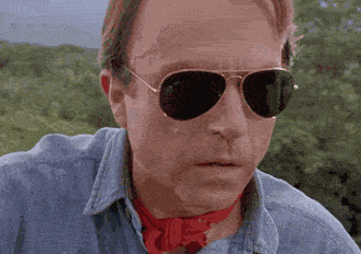 Man wearing aviator sunglasses and red bandana around his neck looking intensely forward outdoors.