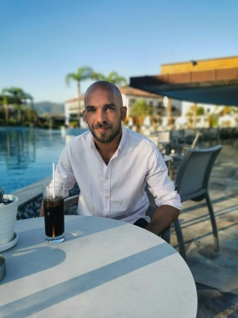 Man in a white shirt sitting at a round table outdoors by a pool with a drink and palm trees in the background.